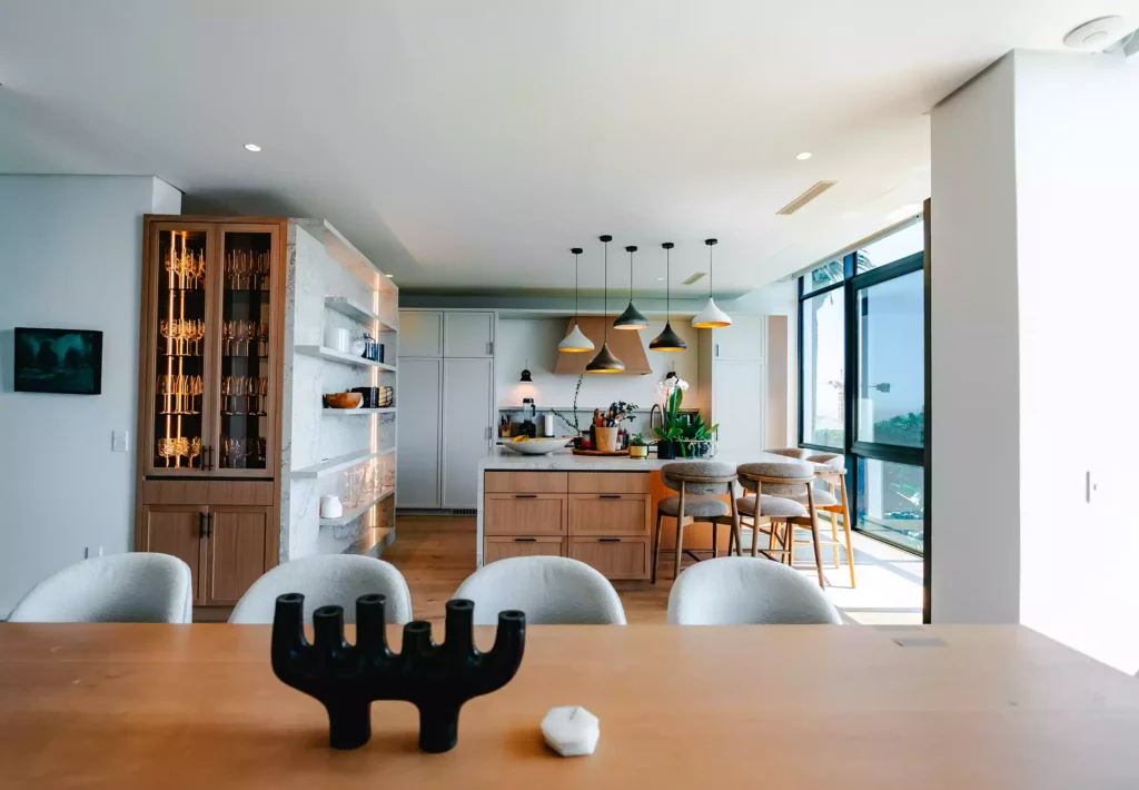 A wide view of a modern open-plan kitchen and dining area taken from across a wooden dining table. The kitchen features a central island with a marble countertop, wood drawers, grey bar stools, and a cluster of hanging pendant lights. On the left stands a tall wood-framed display cabinet with glass doors and internal lighting, sitting next to a built-in marble-look open shelving unit, while large glass windows fill the space with natural light.