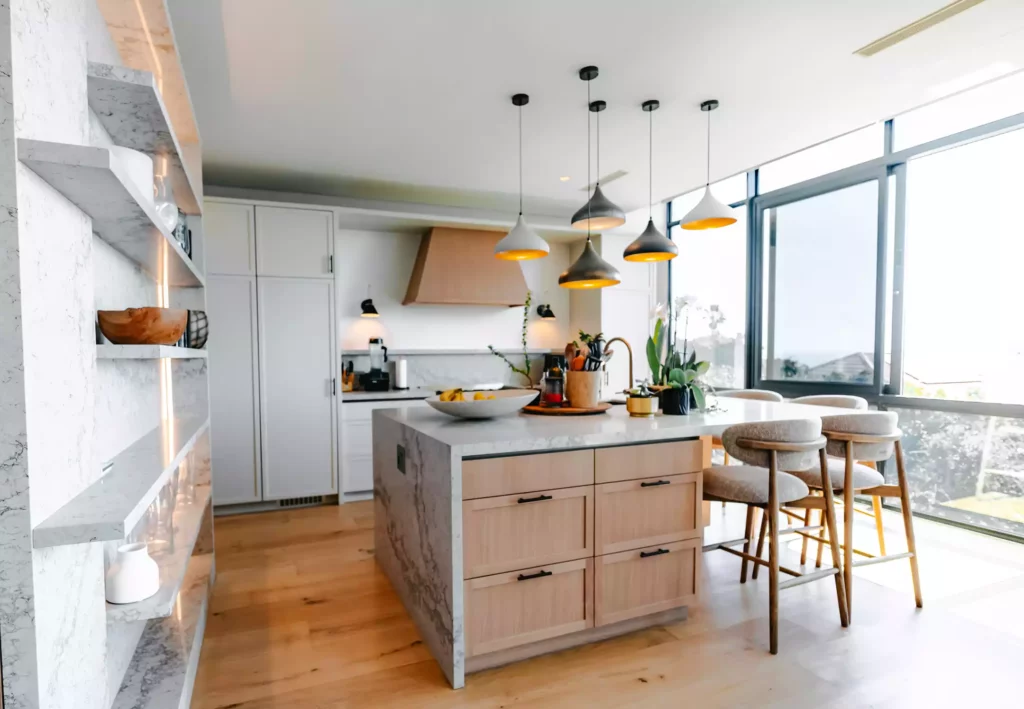A bright, modern kitchen featuring a central island with a white marble waterfall countertop, light wood cabinetry with black handles, and grey upholstered bar stools. A collection of multi-colored pendant lights hangs above the island. The background includes white cabinetry, a light wood extractor hood, and large floor-to-ceiling glass windows offering an outdoor view, while a marble-look open shelving unit stands on the left.