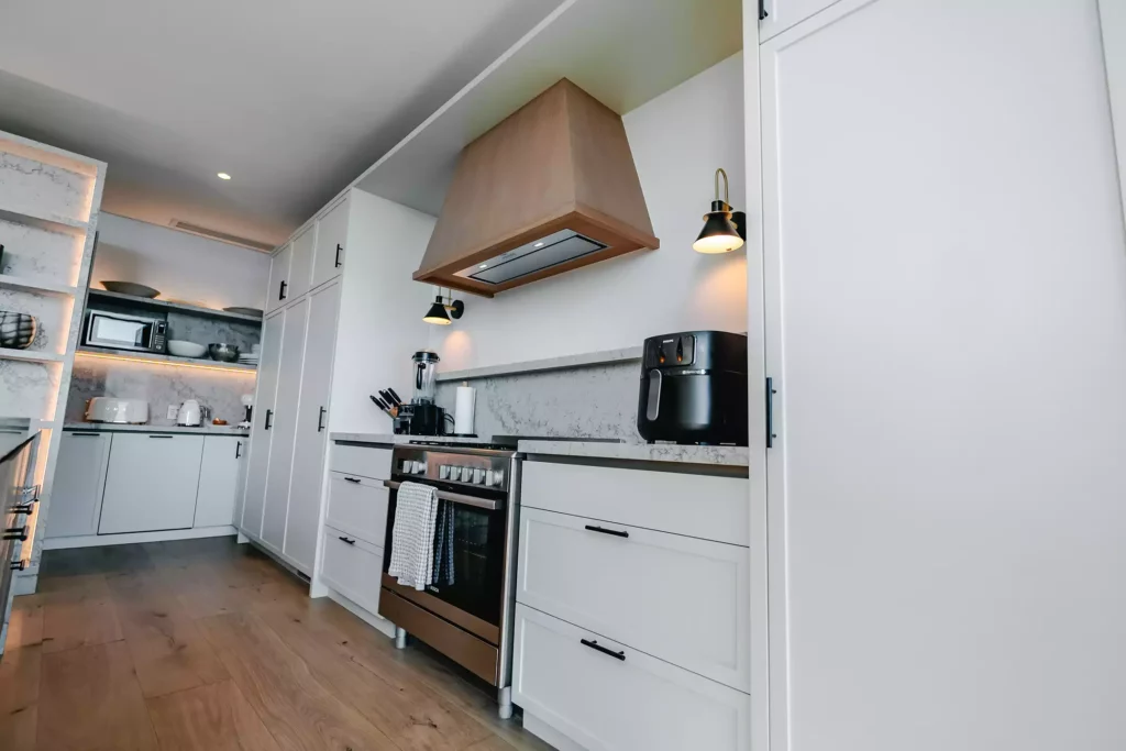A modern kitchen view showcasing sleek white shaker cabinets with black handles, a stainless steel gas range cooker with an oven, a light wood extractor hood, and a marble-look countertop and backsplash. The background opens into a scullery area, while a black air fryer and blender sit on the counter under wall-mounted sconce lights