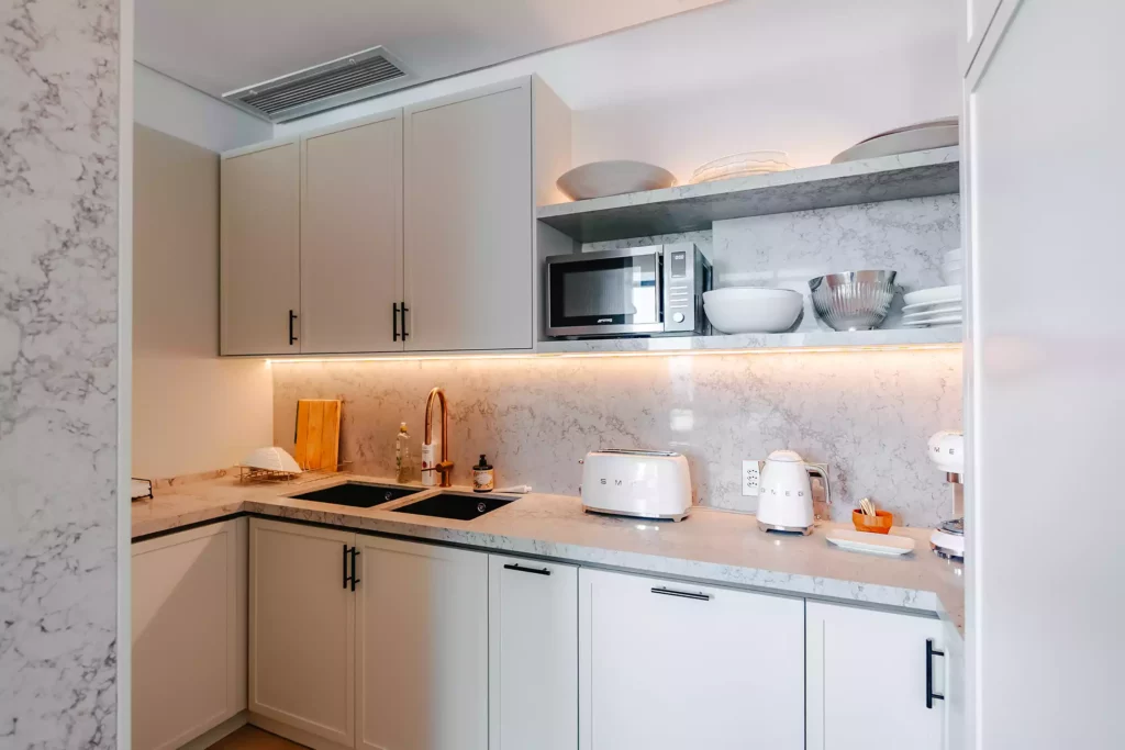A modern kitchen scullery or corner featuring off-white cabinetry with black handles, a white marble-look countertop and matching full-height backsplash, and a gold goose-neck mixer tap over a double black sink. On the right, open marble shelving holds a microwave, mixing bowls, and plates, illuminated by warm under-shelf LED strip lighting.