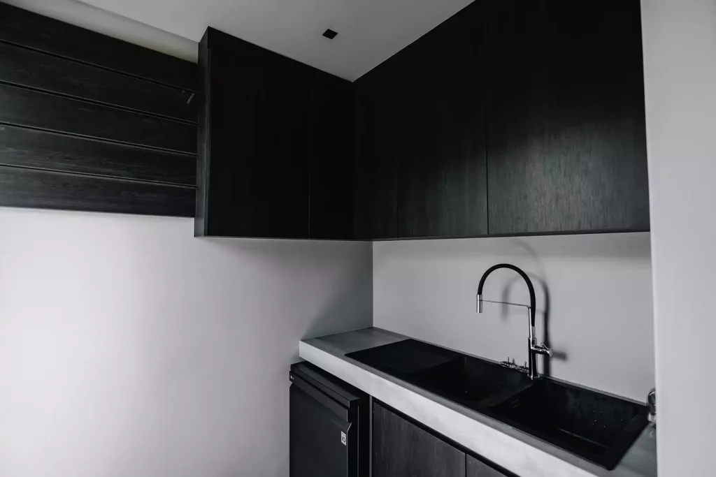 A modern minimalist kitchen scullery featuring matte black upper and lower cabinetry, a light grey concrete-look countertop, and a double black sink. A flexible black and stainless steel goose-neck tap curves over the sink, and a small black under-counter appliance is integrated below.