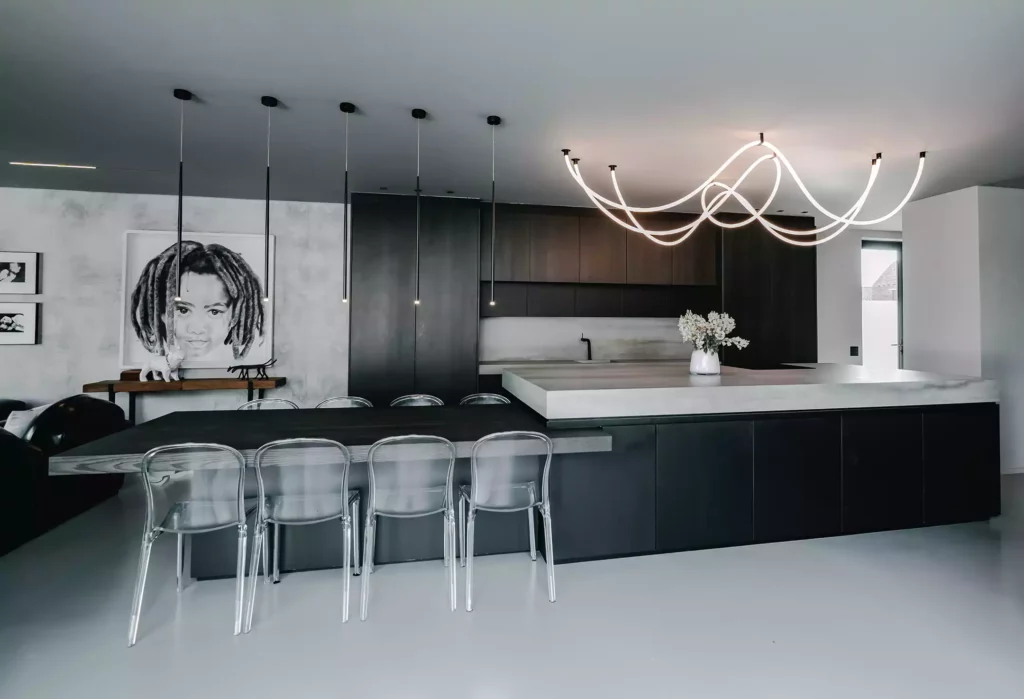 A moody, modern open-plan kitchen and dining area featuring a dark charcoal island with a thick, light grey countertop, illuminated by a large, sculptural looping LED tube chandelier. An attached dark wood dining table is paired with clear acrylic chairs under a row of slim, black pendant lights, while the background shows seamless dark wood cabinetry and a large black-and-white portrait on a textured grey wall.