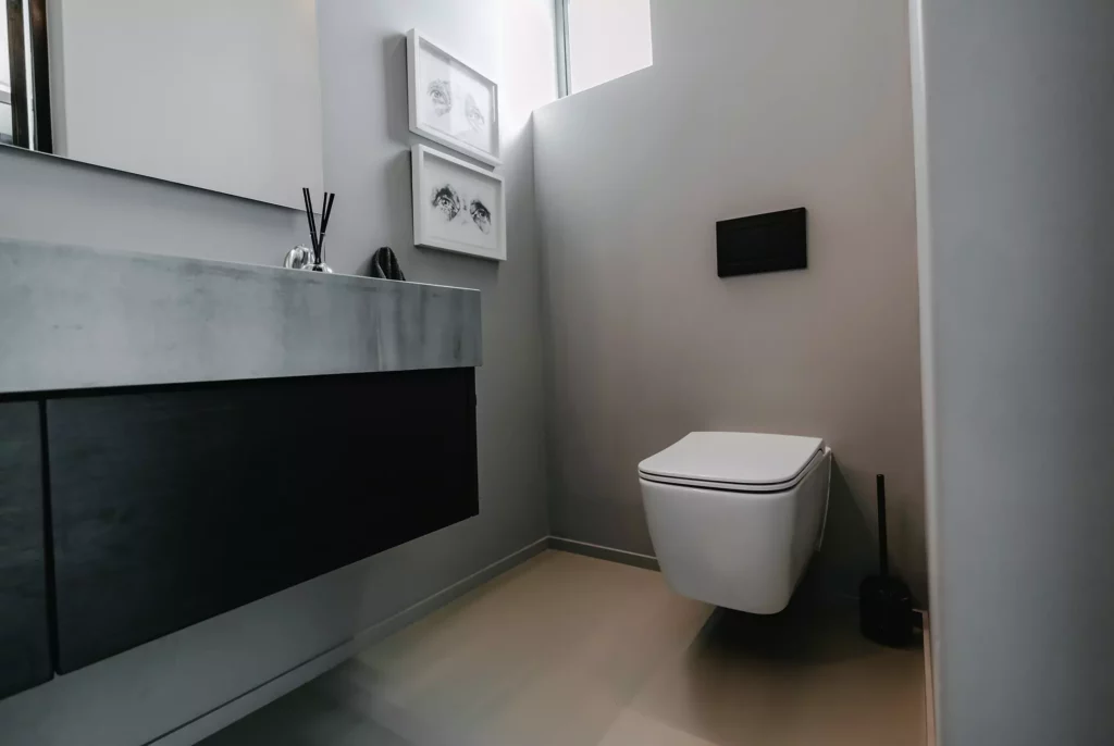 A modern minimalist bathroom featuring a white wall-mounted toilet with a matte black flush plate. On the left sits a floating concrete vanity with sleek dark charcoal drawers underneath. The space is finished with light grey walls, two small framed pieces of art hanging stacked vertically, and a neutral-toned smooth floor.