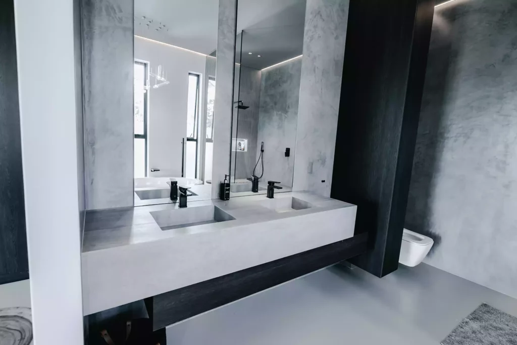 A moody, modern bathroom featuring a long floating concrete double vanity with two integrated rectangular sinks and matte black taps. Tall vertical mirrors sit against textured grey concrete-look walls, reflecting a walk-in shower and freestanding bathtub behind. A dark charcoal wood storage shelf runs beneath the vanity unit, while a white wall-mounted toilet is visible on the right.