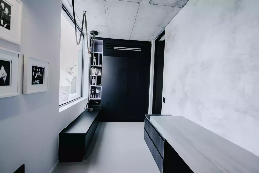 A minimalist corridor or dressing space featuring textured concrete ceilings and a matching light grey wall on the right. The back wall is dominated by sleek, matte black floor-to-ceiling cabinetry and a built-in display shelving unit filled with small decorative figurines. A long, low black bench runs beneath a large window on the left, which is lit by modern, draped pendant bulb lights, while a grey and black console unit stands in the foreground.