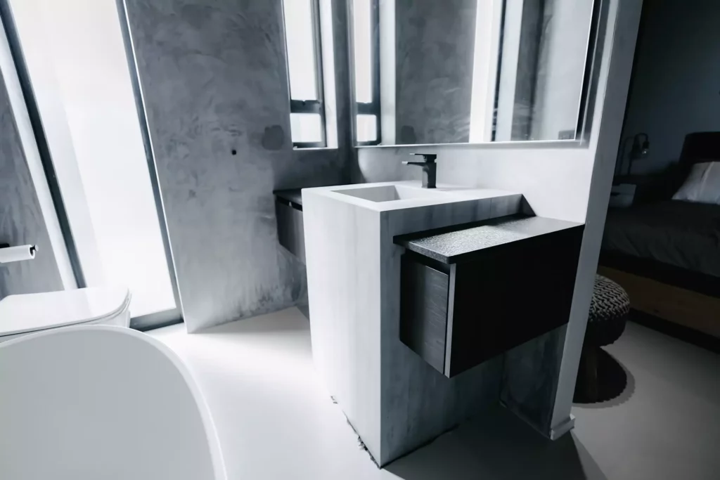 A moody, modern bathroom featuring a unique central concrete-look vanity unit with an integrated square sink, a black tap, and attached floating dark charcoal drawers. The space includes textured grey concrete-look walls, a large mirror, and glimpses of a white freestanding bathtub on the left and an adjacent bedroom on the right.