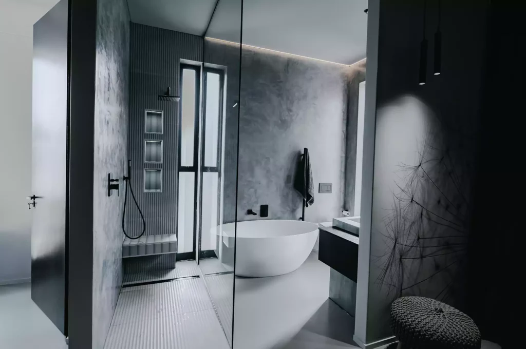 A moody, modern bathroom featuring textured grey concrete-look walls, a freestanding white oval bathtub, and a large glass-enclosed walk-in shower. The shower area includes vertical grey ribbed tiling, integrated storage niches, a rain shower head, and a built-in bench. A black floating vanity sits beside the tub, and a decorative wall with a dandelion graphic is visible in the foreground.