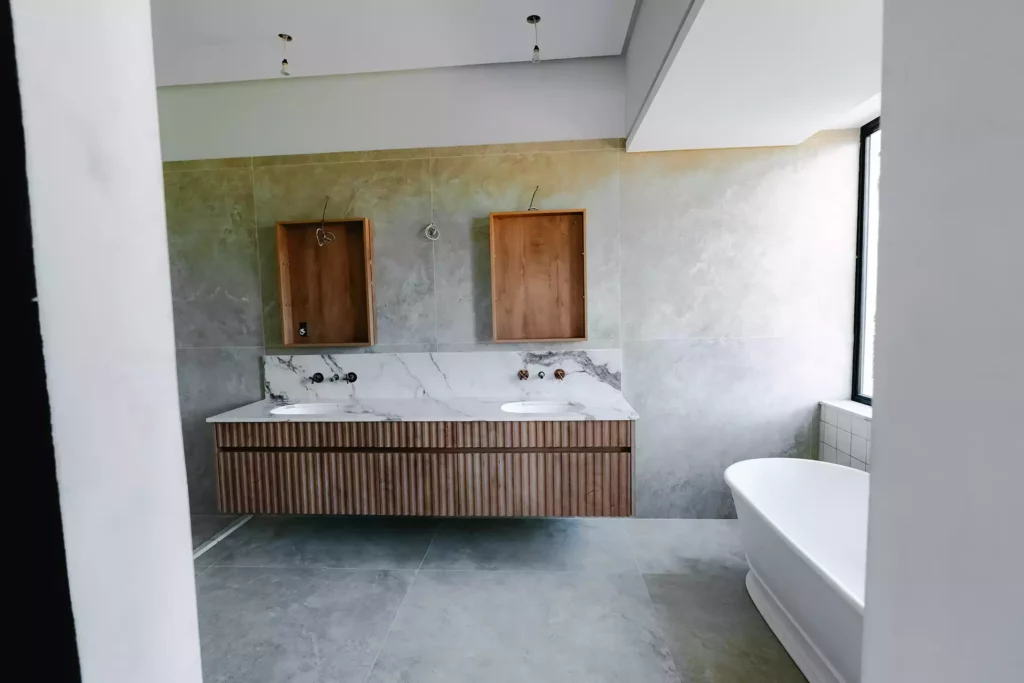 A modern bathroom under construction featuring a floating double vanity with vertical wood slat details, a white marble-look countertop and backsplash with dual integrated sinks, two wooden mirror frames mounted on a textured grey wall, and a freestanding white bathtub on the right.
