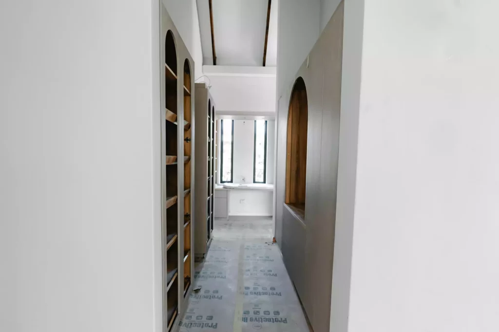 A hallway or walk-in closet area under construction, featuring built-in cabinetry with arched design elements and internal wooden shelving, protective floor covering, and slim vertical windows at the end of the passage.