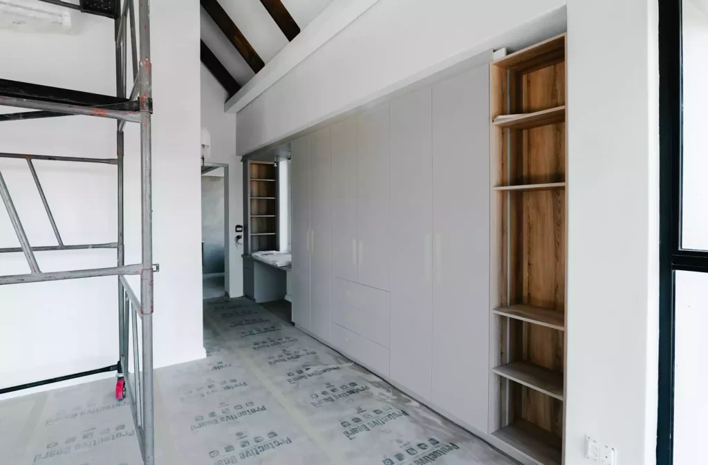 A bedroom or dressing area under construction featuring a large built-in white wardrobe with integrated drawers, an open light wood shelving unit on the right, a desk area in the background hallway, scaffolding on the left, and protective floor covering.
