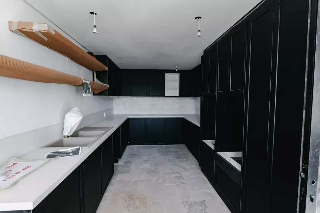 A modern scullery or laundry room under construction, featuring matte black cabinets, white countertops, a double sink, two long floating wooden shelves wrapped in protective cardboard, and grey tiled flooring.