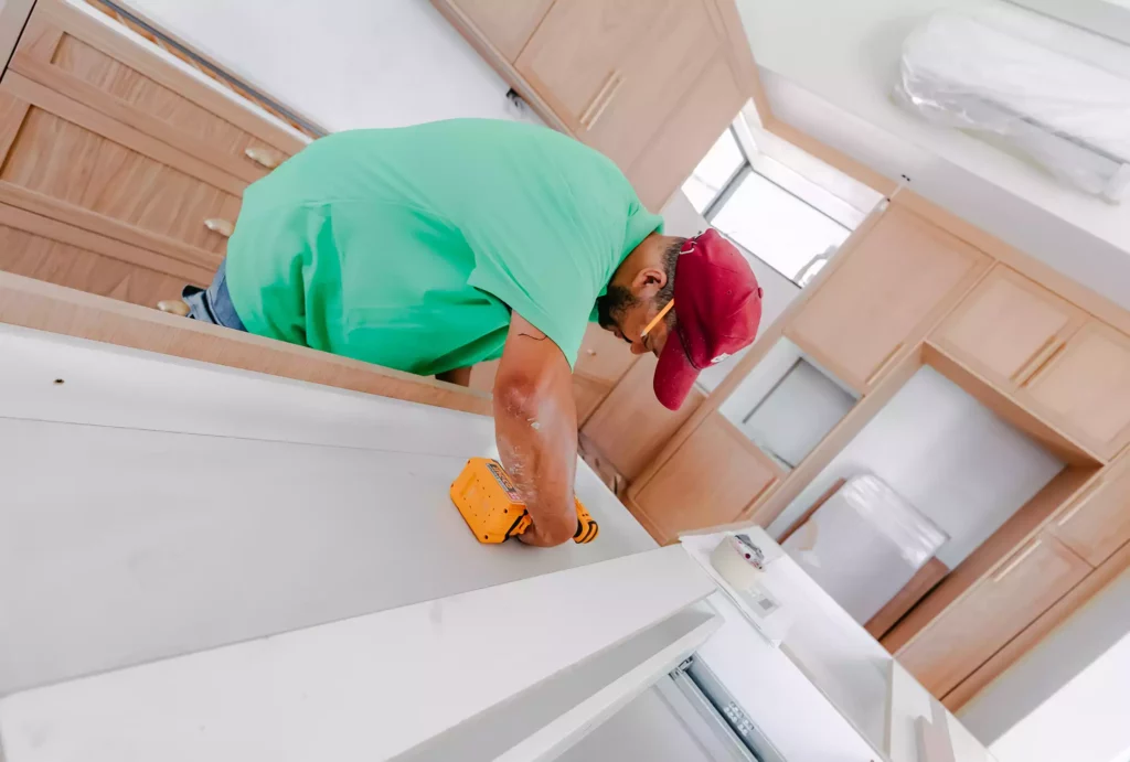 A carpenter wearing a green t-shirt and a red cap uses a yellow cordless drill to assemble light wood kitchen cabinets.