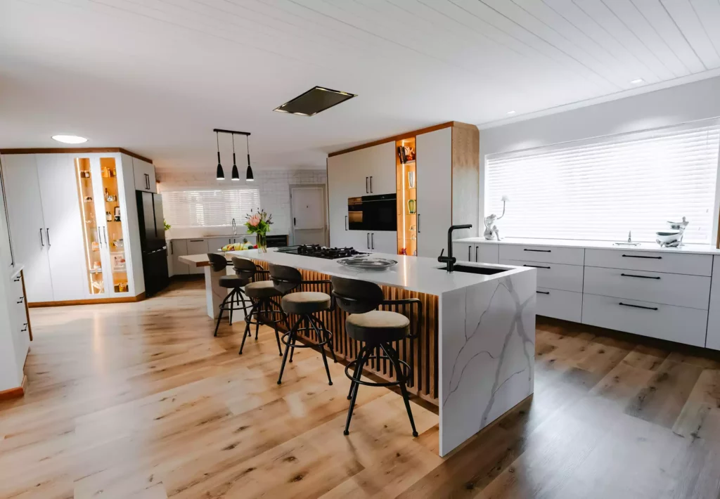 A modern open-plan kitchen featuring a white marble waterfall island lined with wooden slats, black counter stools, white cabinetry, and wood flooring.