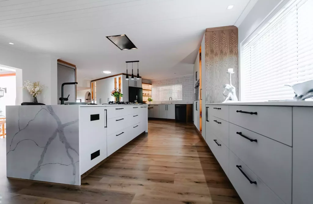A spacious modern kitchen featuring a white marble waterfall island, white cabinetry with black handles, and warm wood flooring.