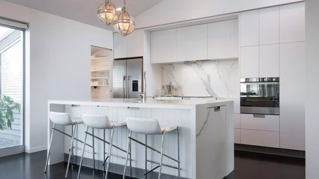 Modern minimalist white kitchen featuring a marble waterfall center island with white leather bar stools, seamless floor-to-ceiling cabinets, stainless steel appliances, and elegant glass globe pendant lights.