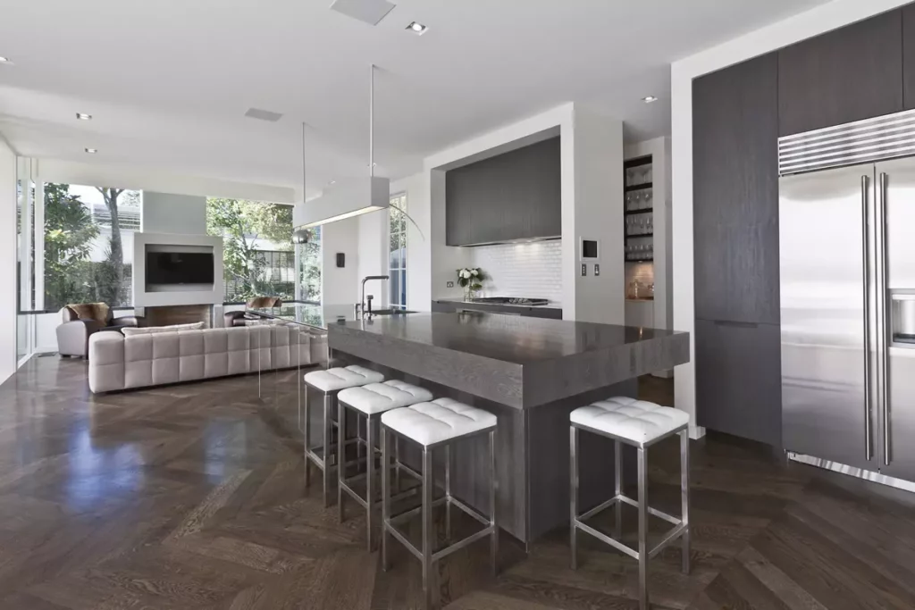 Modern open-plan kitchen and living room featuring a dark wood center island with white tufted bar stools, stainless steel appliances, and herringbone hardwood flooring.