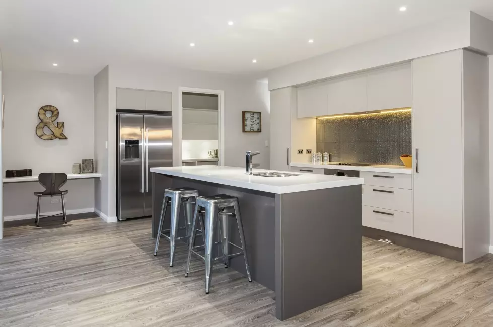 Modern kitchen featuring a grey center island with a white countertop and metal bar stools, stainless steel appliances, a textured grey backsplash, and light wood floors.