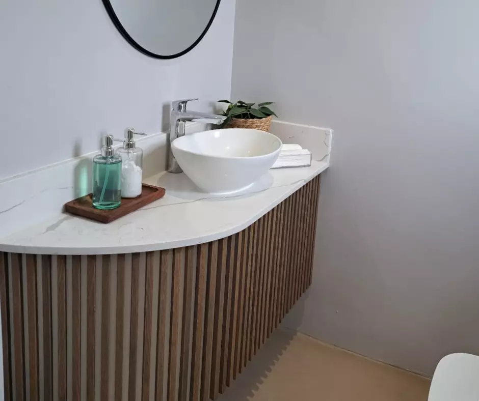 Modern floating bathroom vanity with vertical wooden slats, a curved white marble countertop, a round white vessel sink, and a black-rimmed circular mirror.