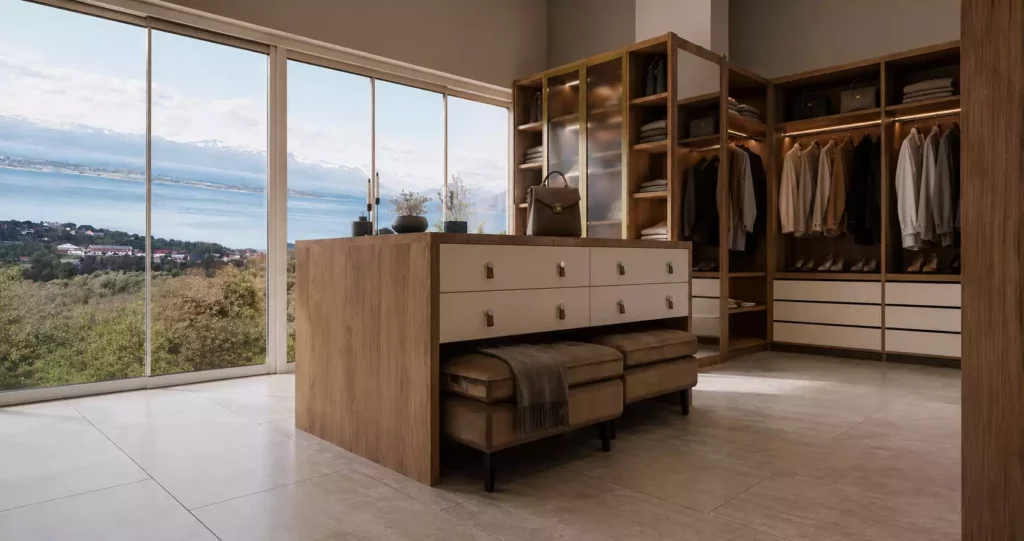 A luxurious walk-in wardrobe with a wooden central island, integrated white drawers, and open garment shelving overlooking a scenic coastal view through large glass windows.