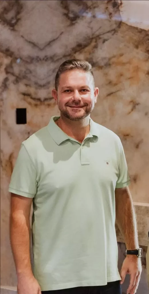 A portrait of a man smiling, wearing a light green polo shirt, positioned in front of a textured marble-patterned wall
