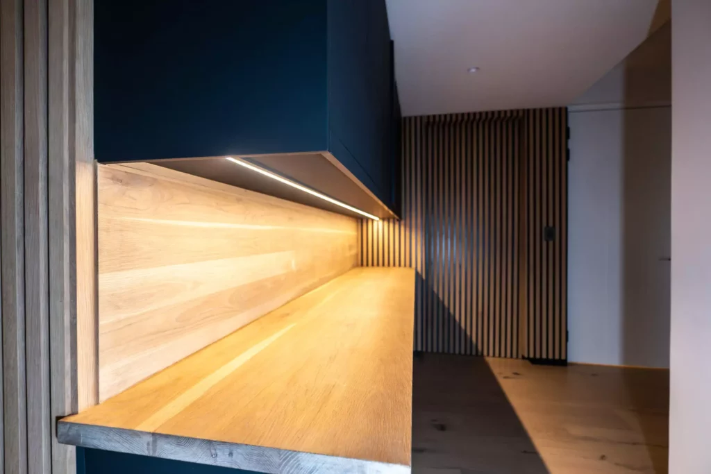 Close-up of a modern kitchen workspace with dark upper cabinets, a warm wooden countertop and backsplash, under-cabinet strip lighting, and adjacent vertical wood slat paneling.