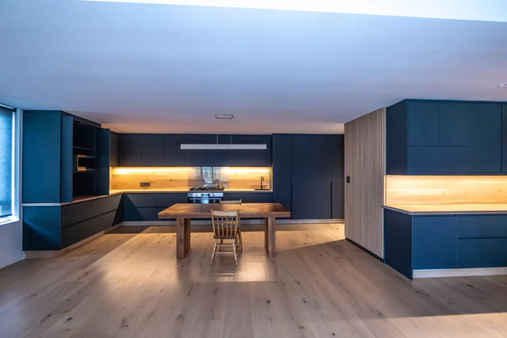 Modern open-plan kitchen featuring dark cabinetry, warm wood flooring, and a central wooden dining table with under-cabinet accent lighting.