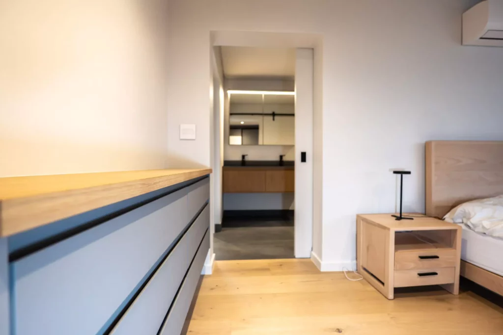 Modern bedroom with a grey chest of drawers, light wood nightstand, and an entrance to the bathroom.
