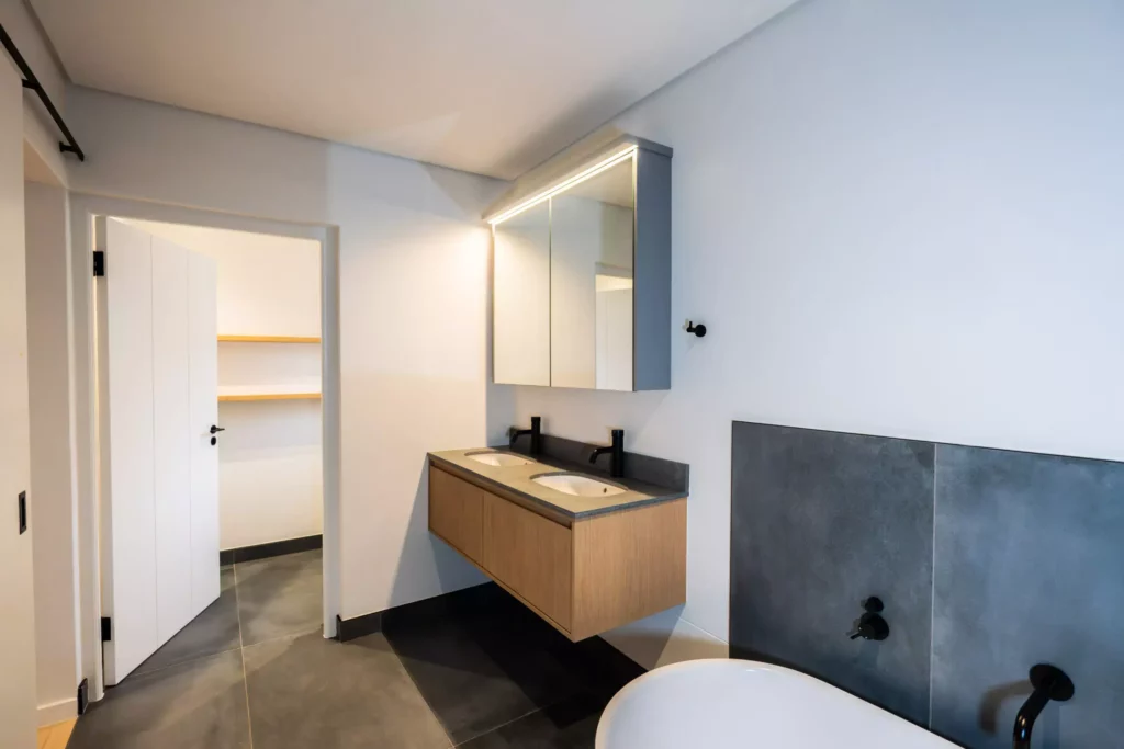 A modern bathroom featuring a floating wooden vanity with a double sink, an illuminated medicine cabinet, a doorway leading to a walk-in storage area with wooden shelves, and a portion of a freestanding bathtub against a dark grey accent wall.