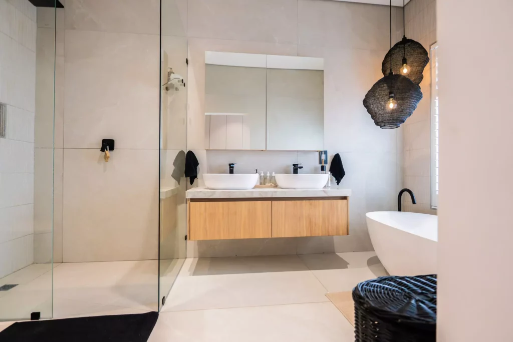 A modern, light-filled bathroom featuring a glass-enclosed shower, a double-sink vanity with a light wood finish, a freestanding white bathtub, and two textured black pendant lights.