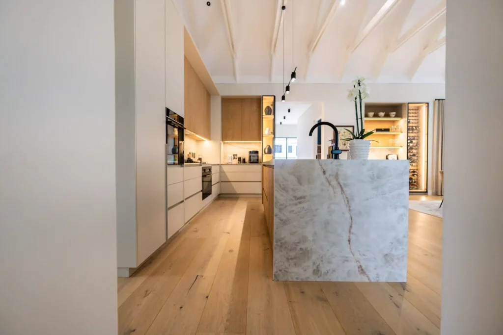 A bright, modern open-plan kitchen featuring a large waterfall marble island with a black faucet, light wood flooring, and high, white vaulted ceilings. The kitchen is equipped with white and warm wood-toned cabinetry, integrated lighting, and a floor-to-ceiling glass display unit on the far wall.