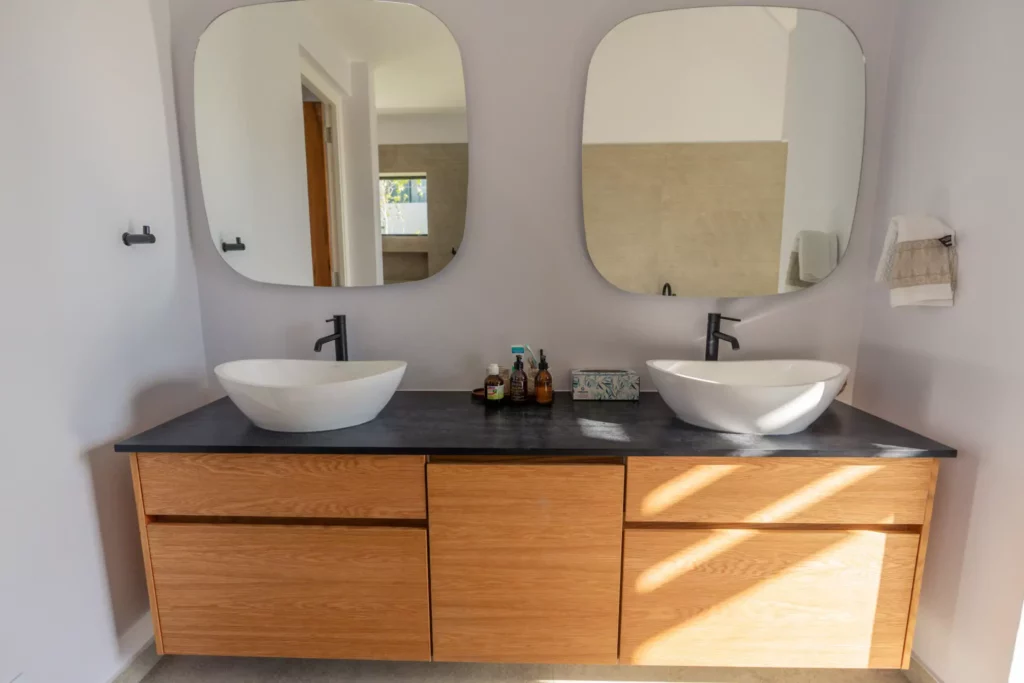 A modern double bathroom vanity with a light oak cabinet, a black countertop, two white vessel sinks with black faucets, and two large, rounded-square mirrors.