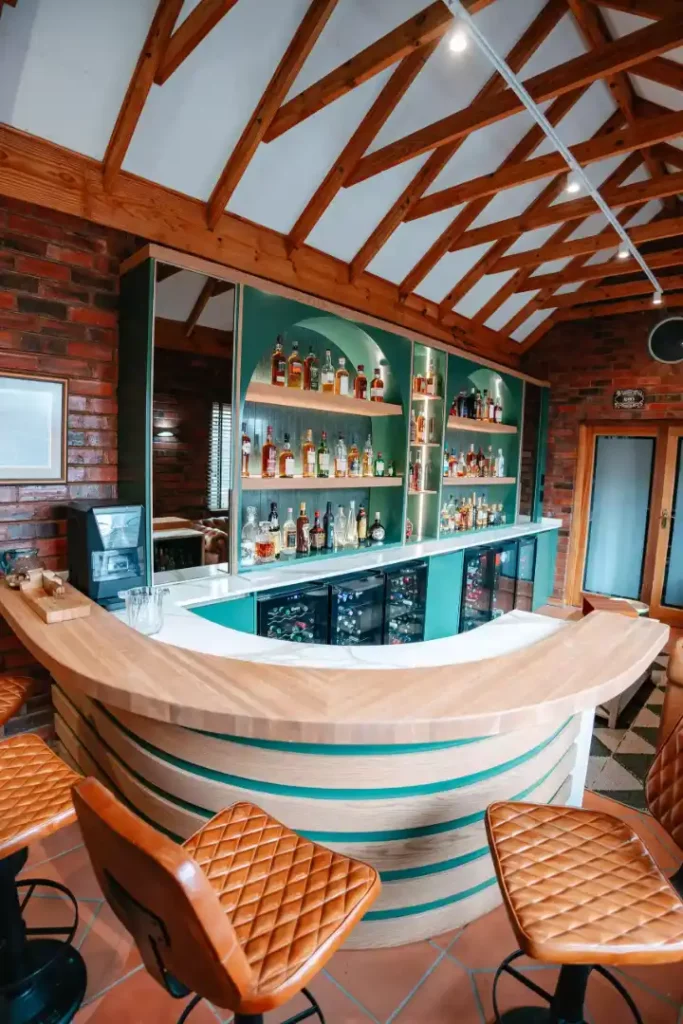 A rustic home bar featuring a curved wooden counter with layered slatted details and green accents, paired with brown quilted leather bar stools. The back bar includes teal cabinetry, illuminated arched shelving displaying various liquor bottles, and integrated glass-front drink fridges under a white marble countertop. The space is set against exposed brick walls beneath a high vaulted ceiling with exposed wooden rafters.