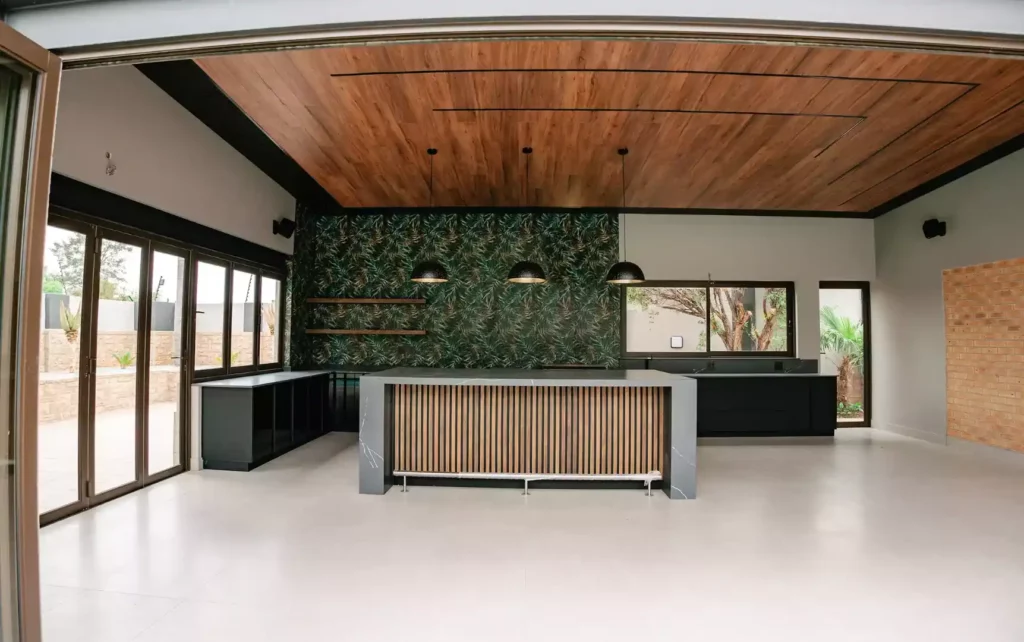 Modern open-plan kitchen and entertainment area viewed through folding doors, featuring a large charcoal marble waterfall island with vertical wood slats, black pendant lights, a tropical leaf-patterned accent wall, and a wood-paneled ceiling.