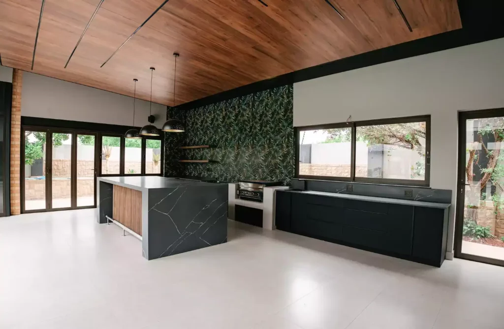 Modern open-plan kitchen and entertainment area featuring a large charcoal marble waterfall island with vertical wood slat details, matte black lower cabinets, a built-in braai station, a prominent tropical leaf-patterned accent wall, and a warm wood-paneled ceiling.