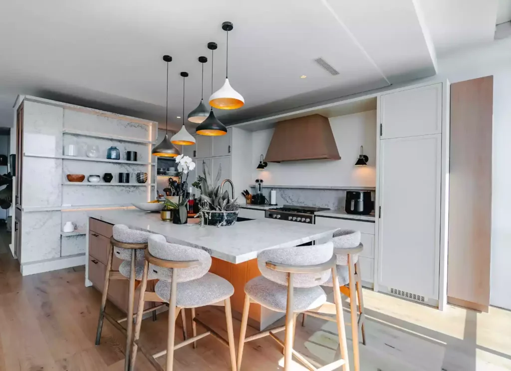 A bright, modern open-plan kitchen featuring a large central island with a white marble countertop and integrated wood cabinetry, paired with grey upholstered bar stools with wooden frames. Above the island hangs a cluster of multi-colored pendant lights with gold interiors. The back wall includes sleek white cabinets, a gas range cooker beneath a light wood extractor hood, and a prominent built-in open shelving display unit with a marble-look backing on the left.