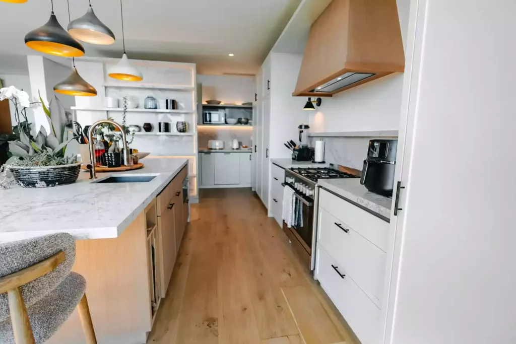 A bright, modern open-plan kitchen featuring a light wood central island with a white marble countertop, a built-in sink, and a gold goose-neck mixer tap. On the right, sleek white cabinets with black handles frame a stainless steel gas range cooker with a light wood extractor hood above it. The space includes light wood flooring, a cluster of multi-colored pendant lights hanging over the island, and a background view of a scullery with open shelving.