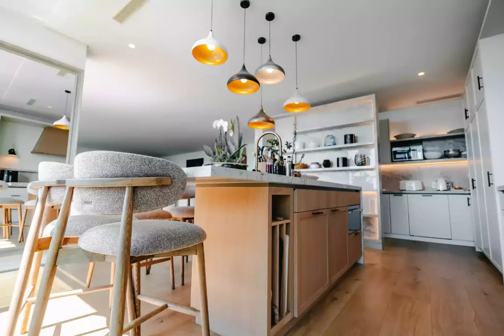 A bright, modern open-plan kitchen featuring a light wood central island with a white marble countertop and integrated shelving. Grey upholstered bar stools with wooden frames are positioned at the counter, beneath a cluster of multi-colored pendant lights with gold interiors. The background shows white cabinetry, a built-in scullery area with open marble shelves, and light wood flooring.