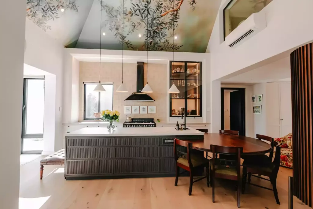 Modern kitchen with a large dark island, wooden dining table, and mural ceiling.