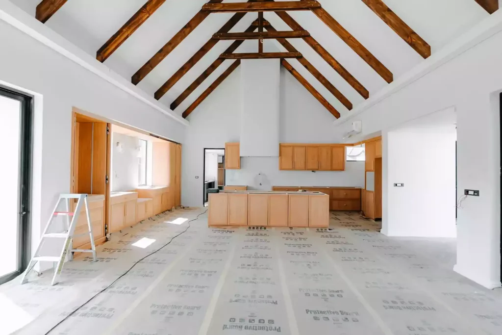 A wide-angle view of a large kitchen under construction, featuring light wood cabinets, an unfinished central island carcass, a high vaulted ceiling with exposed wooden A-frame trusses, protective floor covering, and a stepladder on the left.