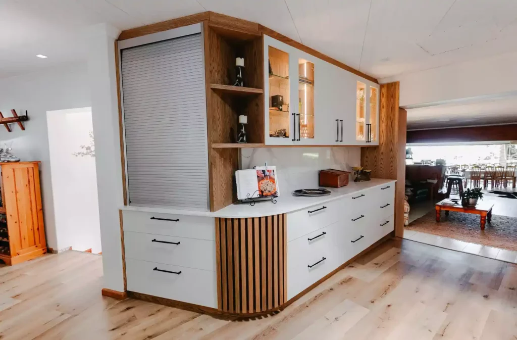 A modern white and wood kitchen coffee station with a silver roller shutter door, illuminated glass-front upper cabinets, and white lower drawers with black handles.