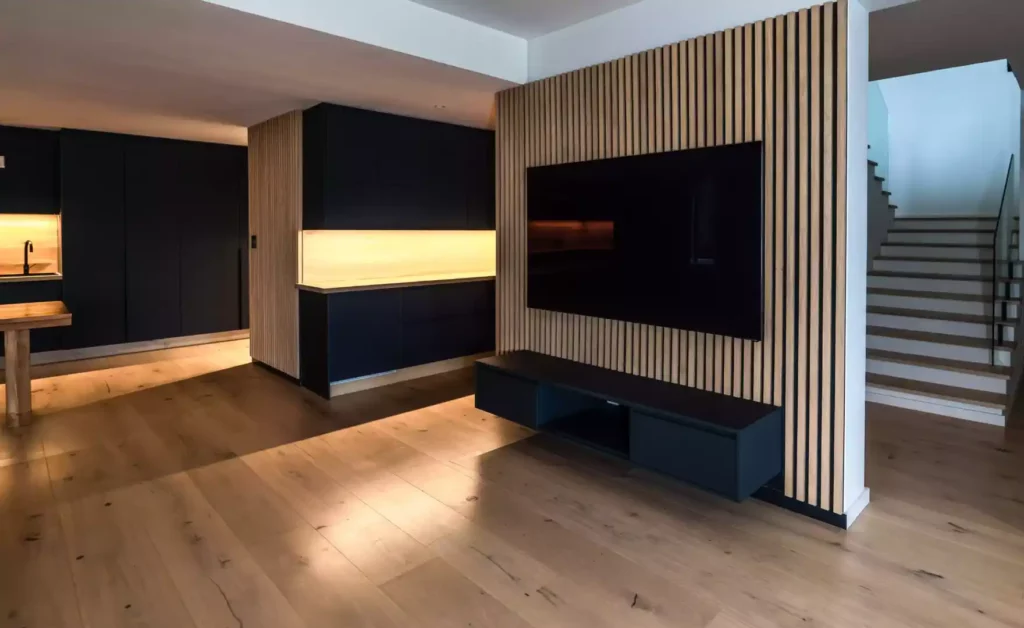 Modern open-plan living area featuring a wall-mounted TV on a wood-slat paneled wall, a black floating TV console, dark kitchen cabinetry with under-cabinet lighting, and wooden flooring leading to a staircase.