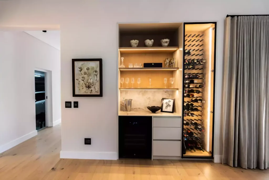 A modern, built-in home bar area featuring illuminated open wooden shelving for glassware, a wine cooler, three storage drawers, and an adjacent vertical wine rack with integrated lighting.