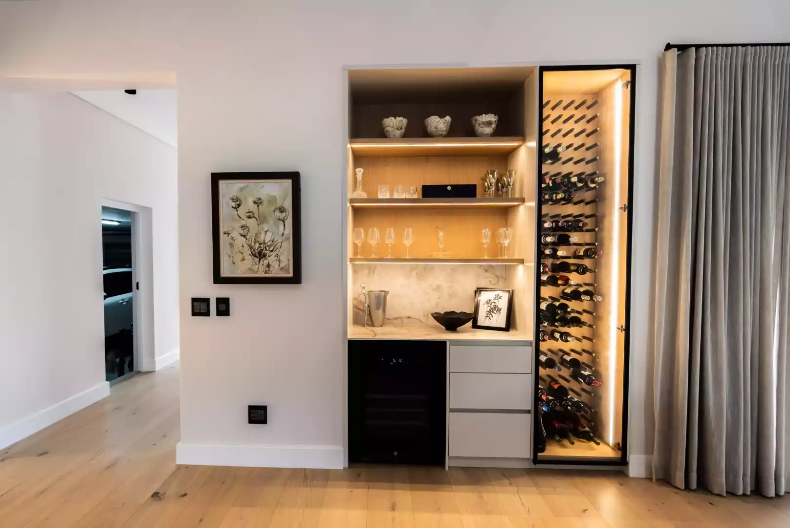 A modern, built-in home bar area featuring illuminated open wooden shelving for glassware, a wine cooler, three storage drawers, and an adjacent vertical wine rack with integrated lighting.