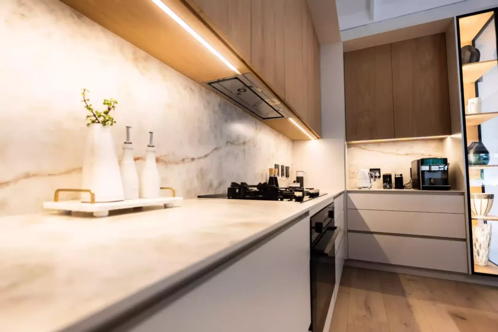 A modern, high-end kitchen featuring minimalist white lower cabinets, warm wood upper cabinetry with integrated under-cabinet lighting, and a luxurious marble-look countertop and backsplash, complemented by an illuminated glass display cabinet and professional appliances