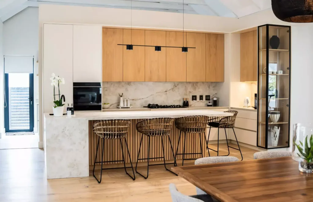 A modern, luxury kitchen featuring extensive warm wood cabinetry, a large kitchen island with a marble countertop, an integrated glass-fronted display section, and wood-slat detailing. The space is accented by three distinctive pendant lights, views of mountainous terrain through large windows, and a doorway with black framing leading to a staircase.