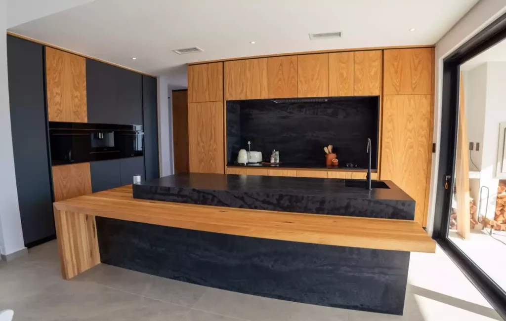A modern open-plan kitchen featuring wood panel cabinetry, a matte black accent wall with integrated appliances, and a prominent black stone island with an attached floating light wood breakfast bar.