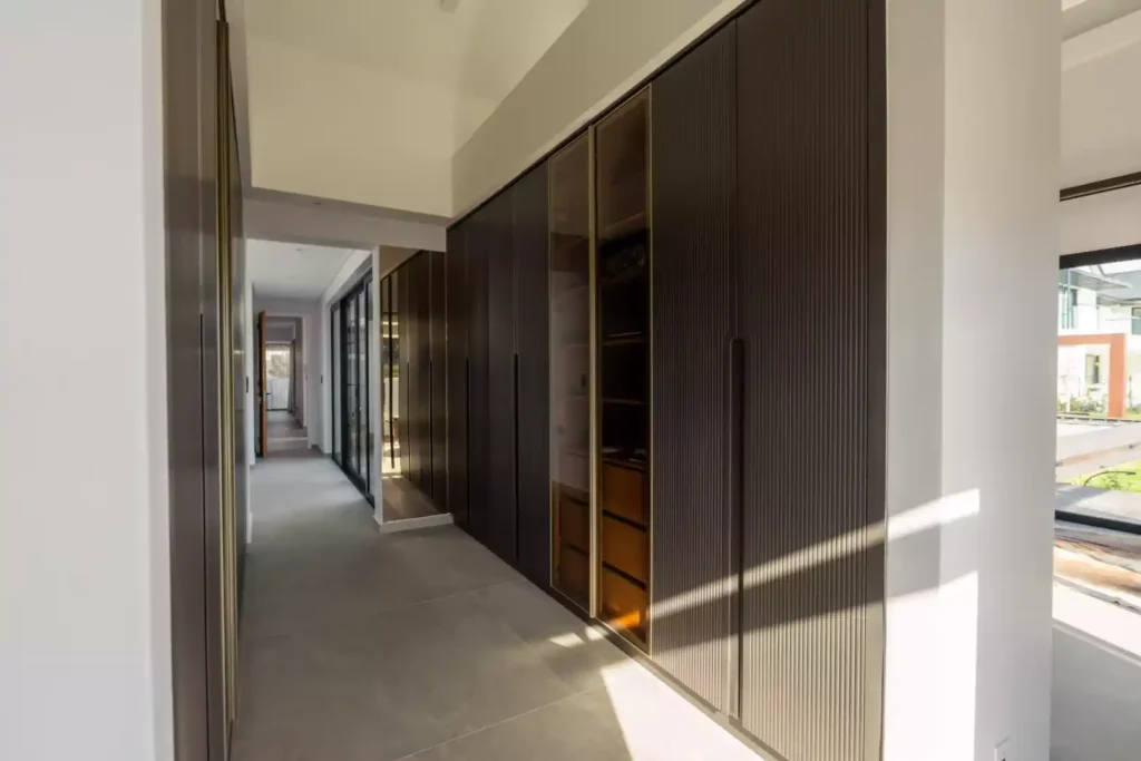 A modern hallway lined with floor-to-ceiling dark grey fluted wardrobes, featuring a central section with glass-front doors revealing illuminated wooden shelves.