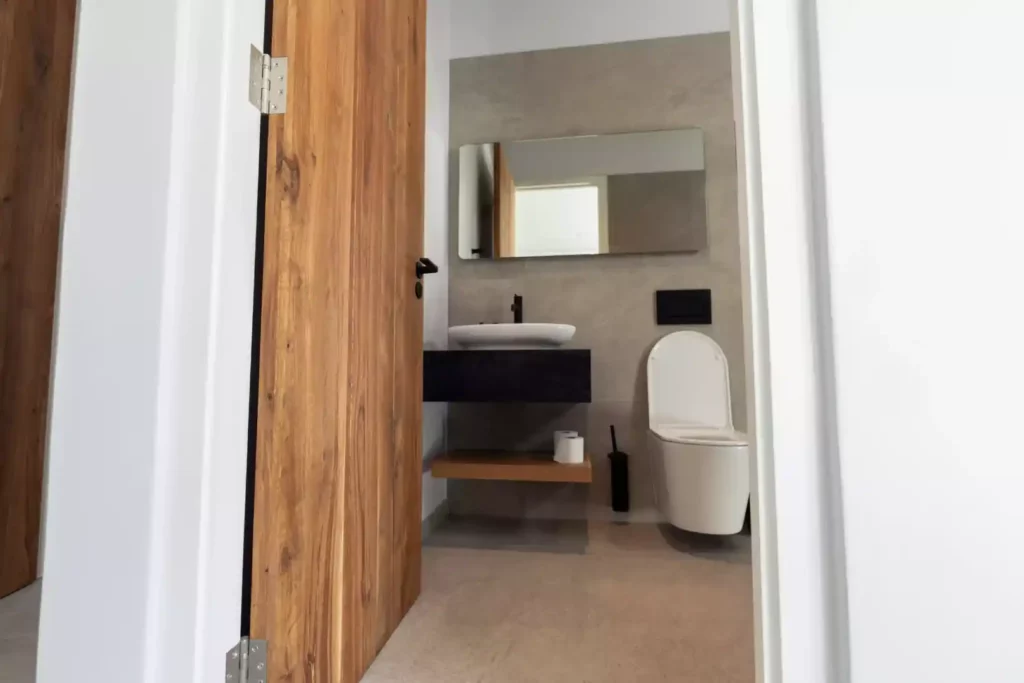 A modern bathroom view through a wooden door, featuring a floating black vanity with a white vessel sink, a rectangular mirror, and a wall-hung toilet against grey tiled walls