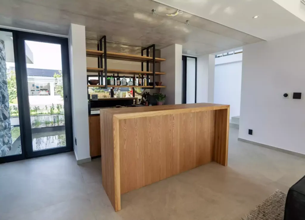 A modern home bar with a light oak waterfall counter, industrial open shelving displaying liquor bottles, and a concrete ceiling.