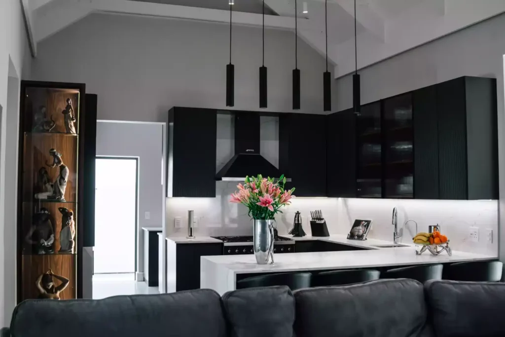 Modern open-plan kitchen featuring black cabinetry, a white marble countertop and backsplash, a central breakfast island with bar stools, and a tall glass display cabinet on the left filled with figurines.